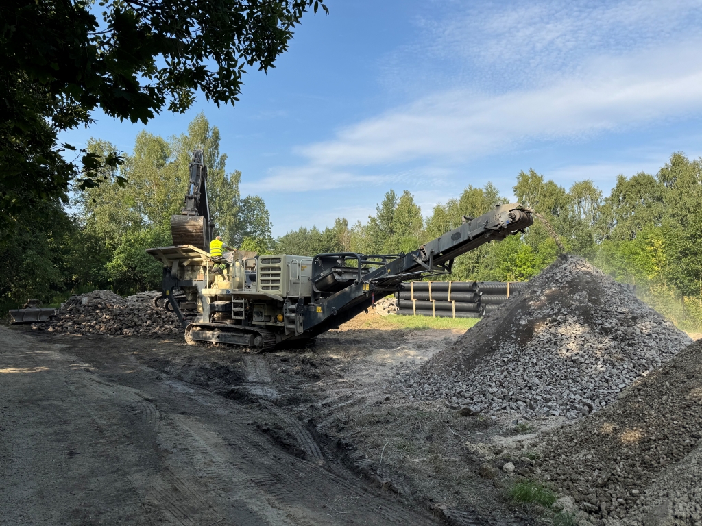 Construction machine with conveyor belt piling gravel in a clearing with trees and blue sky.