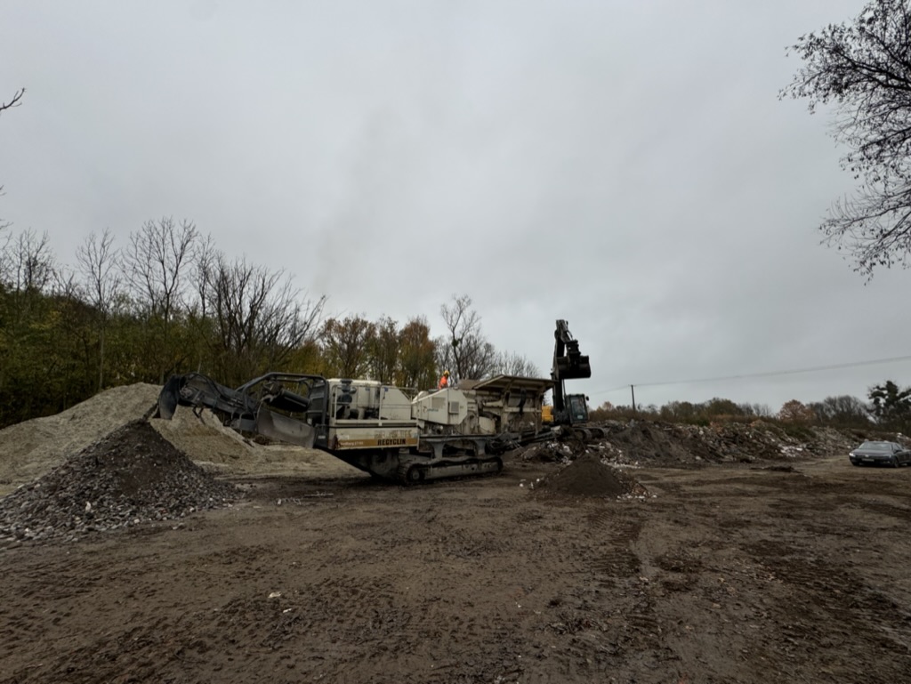 Industrial machine on muddy ground at a construction or recycling site with piles of dirt and trees in background.