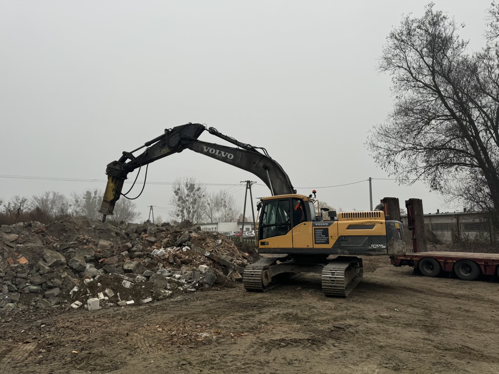 Yellow Volvo excavator with breaker attachment working on rubble pile at a construction site under gray sky.