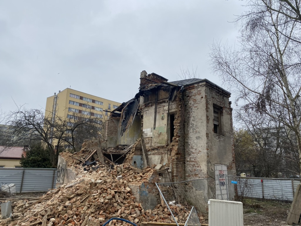 Partially collapsed brick building with rubble pile in front under cloudy sky.