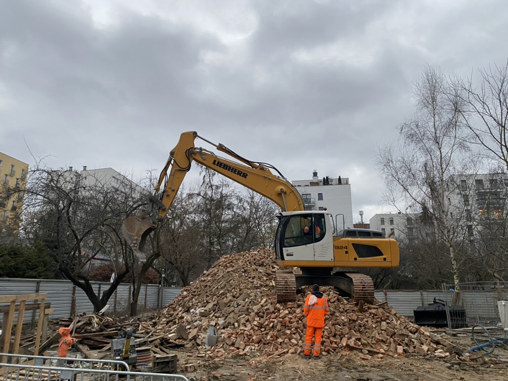 Construction worker in orange stands by a yellow excavator on a large pile of rubble at a site.