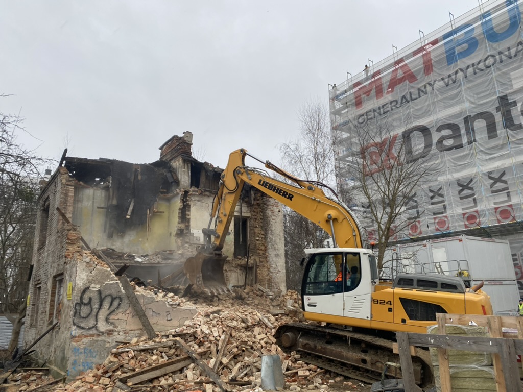 Yellow excavator demolishing old brick building with rubble and scaffolding nearby.