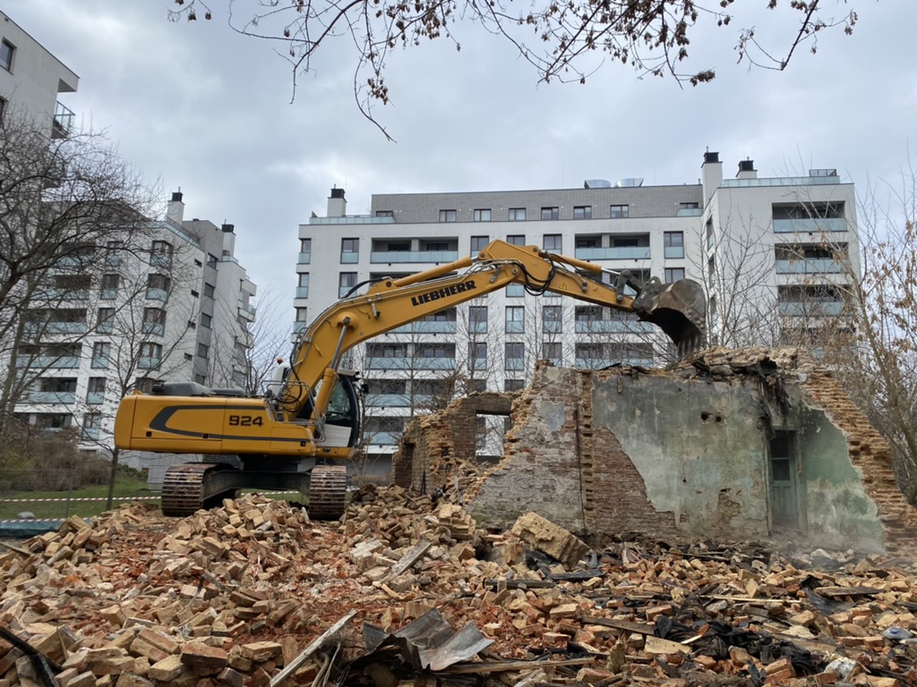 Yellow excavator demolishing old brick building rubble, new apartment buildings in background.