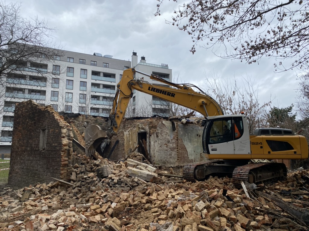 Yellow excavator demolishing an old brick building with rubble scattered around and a modern apartment in background.