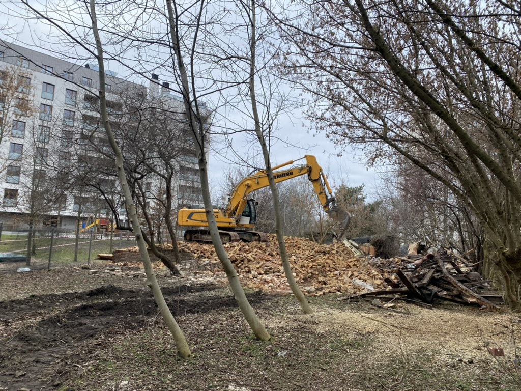 Yellow excavator on a pile of bricks clearing debris near leafless trees and modern buildings.