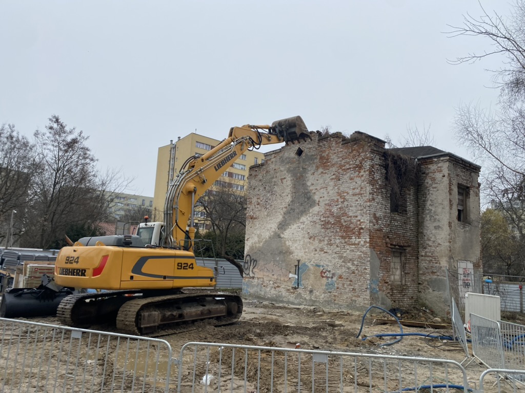 Yellow excavator demolishing old brick building at construction site on a cloudy day.