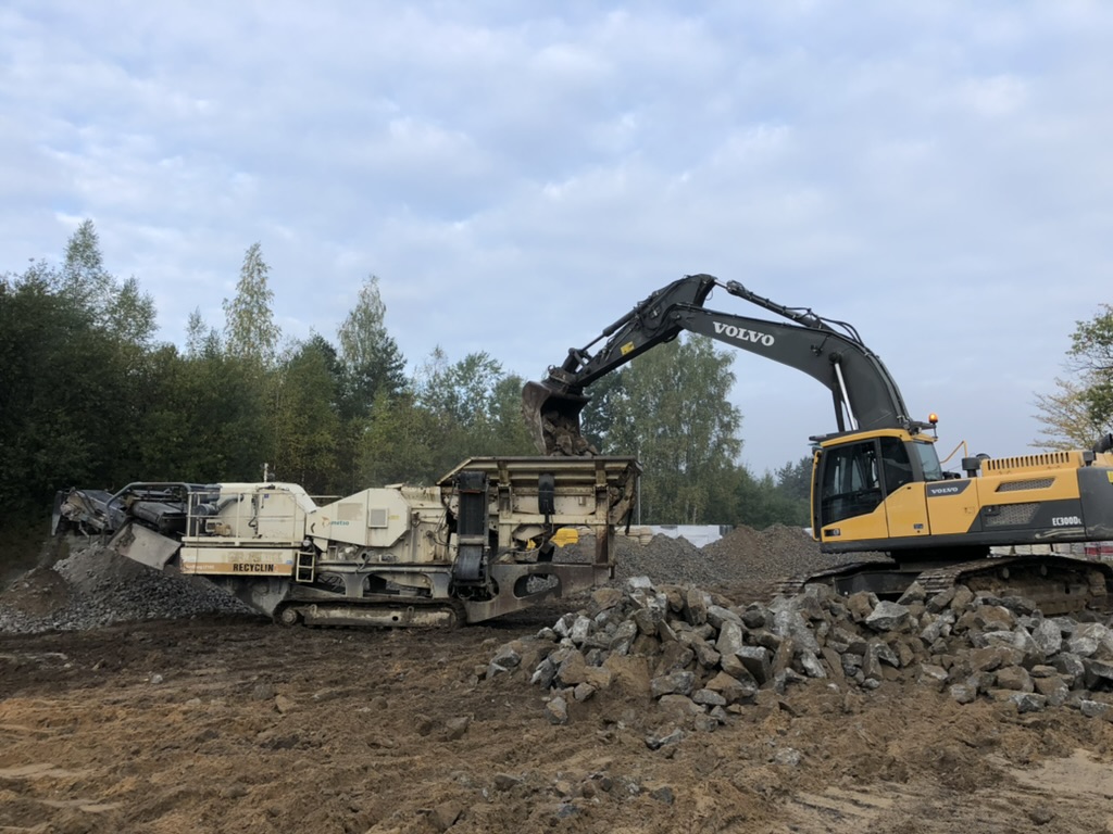 Yellow Volvo excavator loads rocks into white recycling machine on muddy construction site.