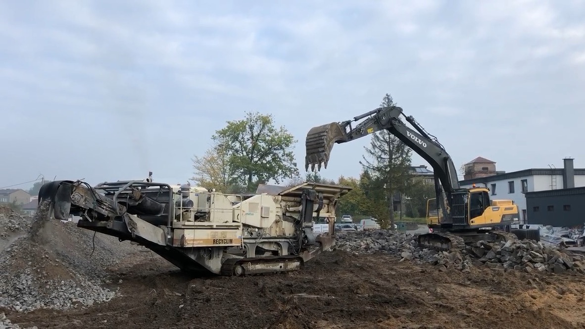 Excavator loading debris into a mobile crusher at a construction site with buildings and trees in background.