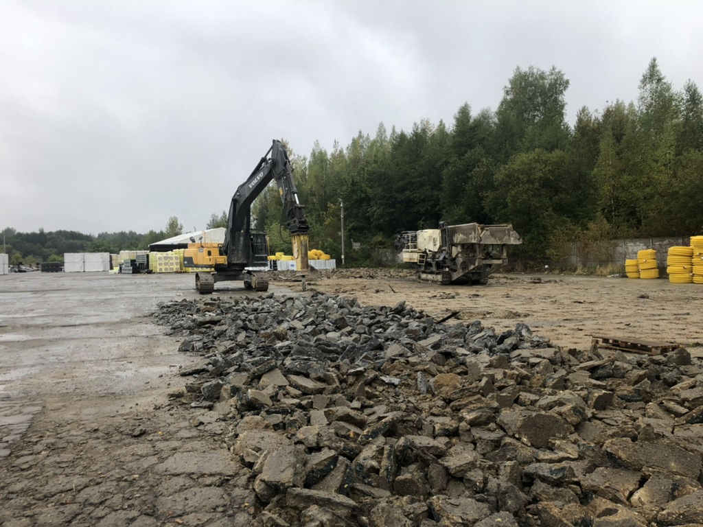 Construction site with excavator breaking asphalt and piles of rubble on the ground near trees.