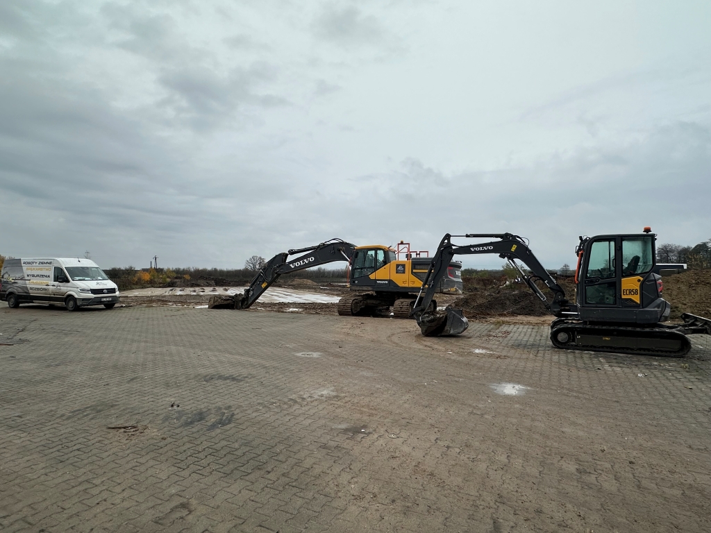 Two Volvo excavators parked on a paved area near a construction site with a white van to the left.