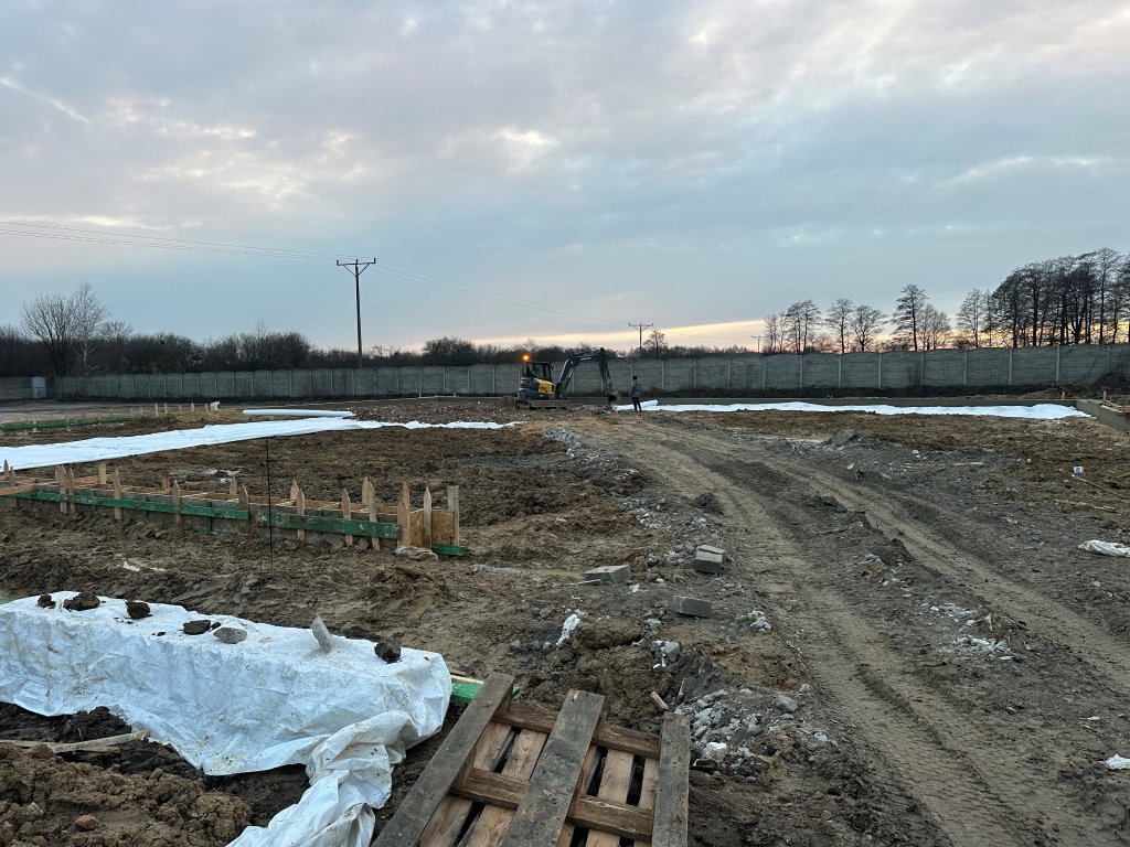 Construction site with excavator, dirt paths, white ground covers, and a person near machinery at dusk.