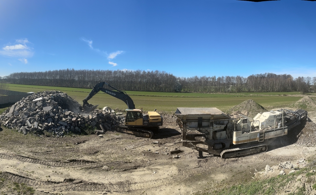 Excavator loading rubble into crusher machine at a construction recycling site on a sunny day.
