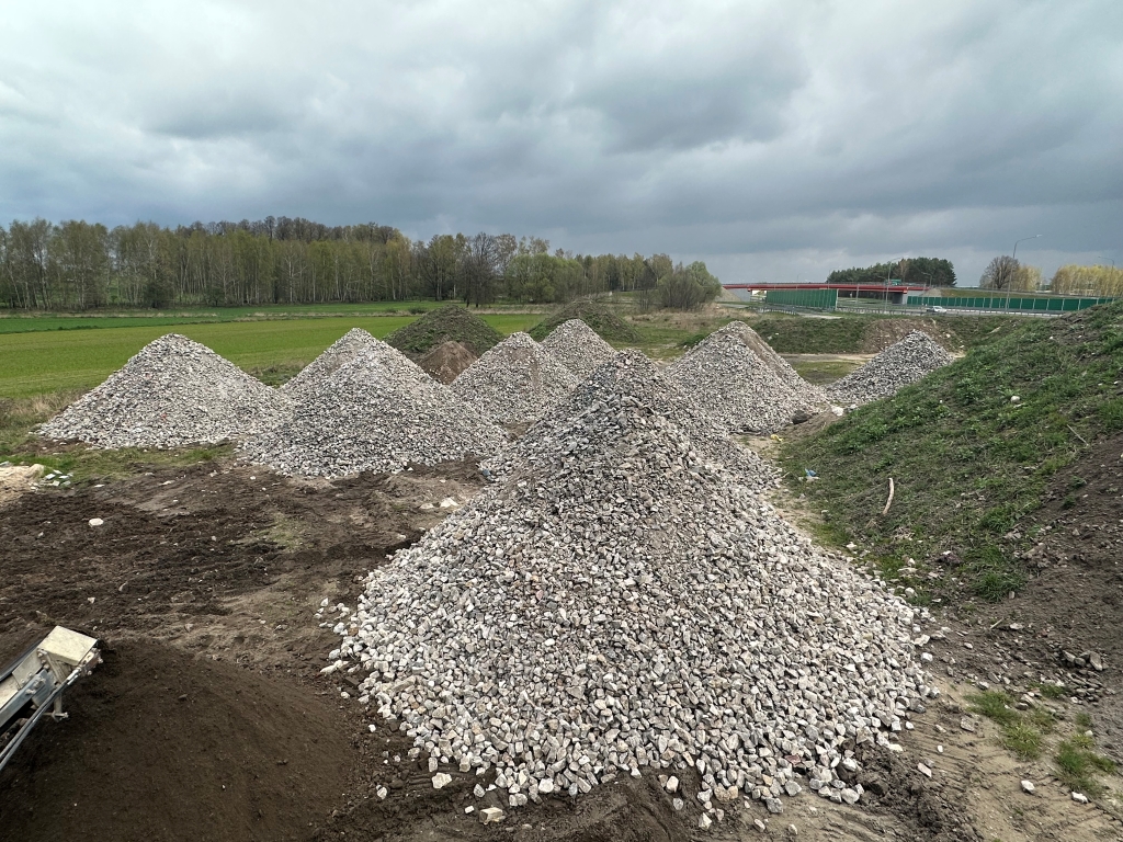 Multiple large piles of gravel and dirt arranged outdoors under a cloudy sky near a grassy field.