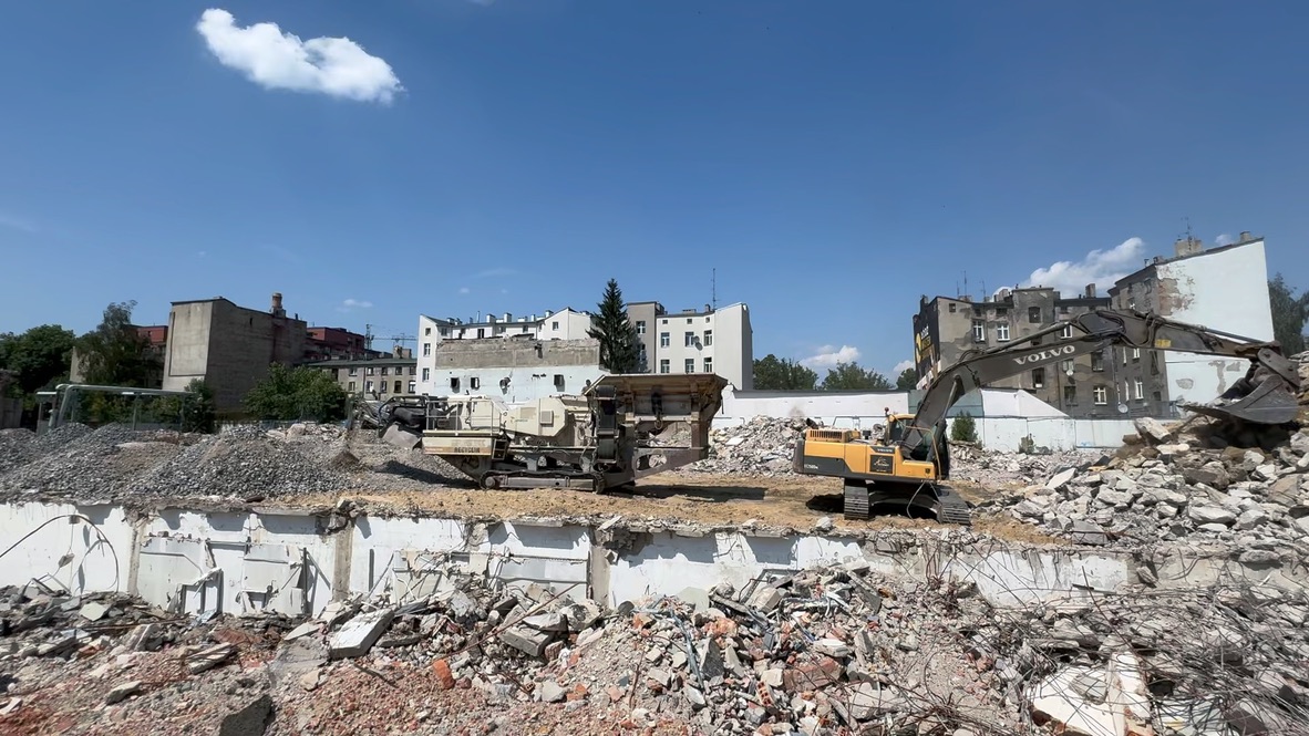 Construction site with excavator and machinery amid rubble and damaged buildings under clear sky.