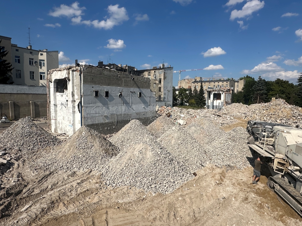 Construction site with rubble piles, partially demolished buildings, and a man near heavy equipment.