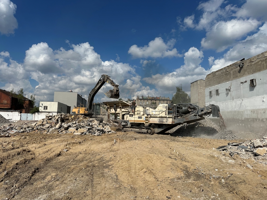 Excavator loading rubble into crusher machine on a construction site with cloudy blue sky.