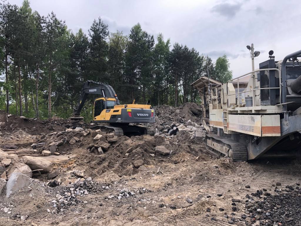 Yellow Volvo excavator and heavy recycling equipment at a rubble-filled construction site near trees.