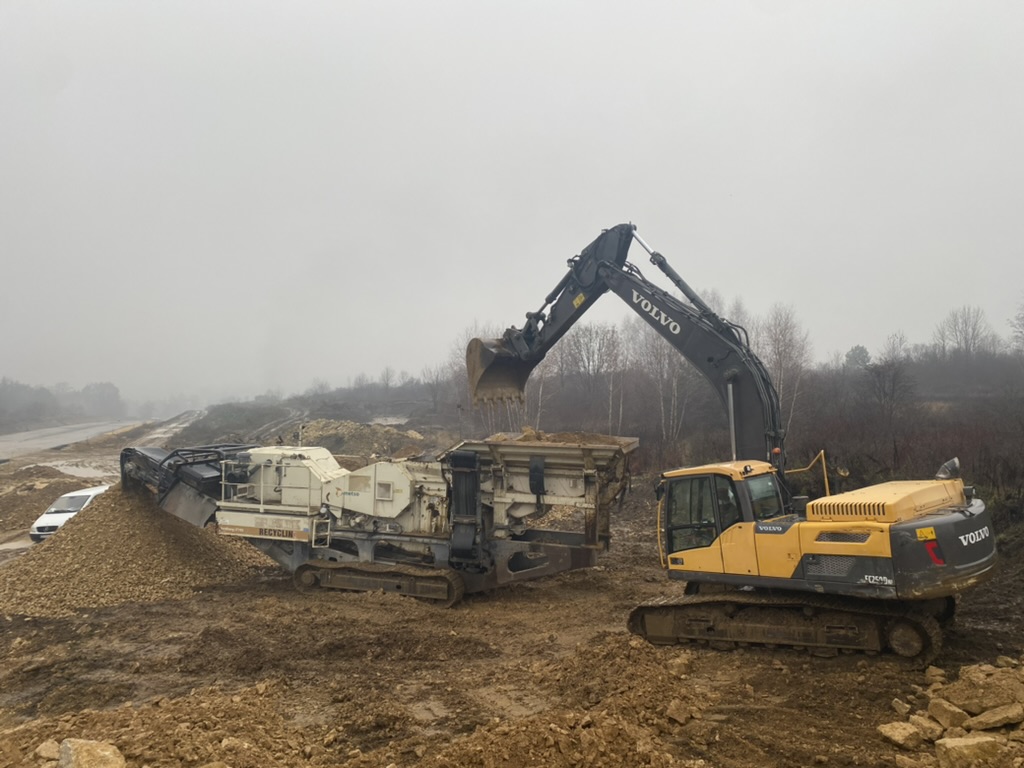 Yellow Volvo excavator loading dirt into a machine at a muddy construction site with overcast sky.