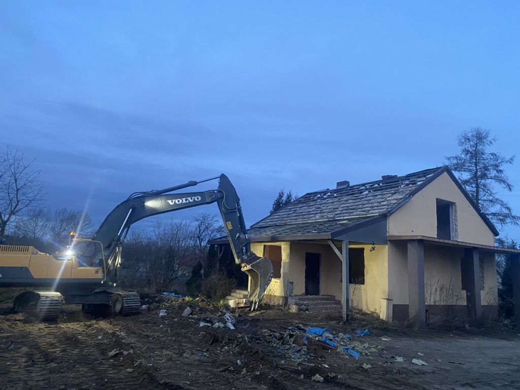 Excavator with lights on next to a partially demolished house under a blue evening sky.