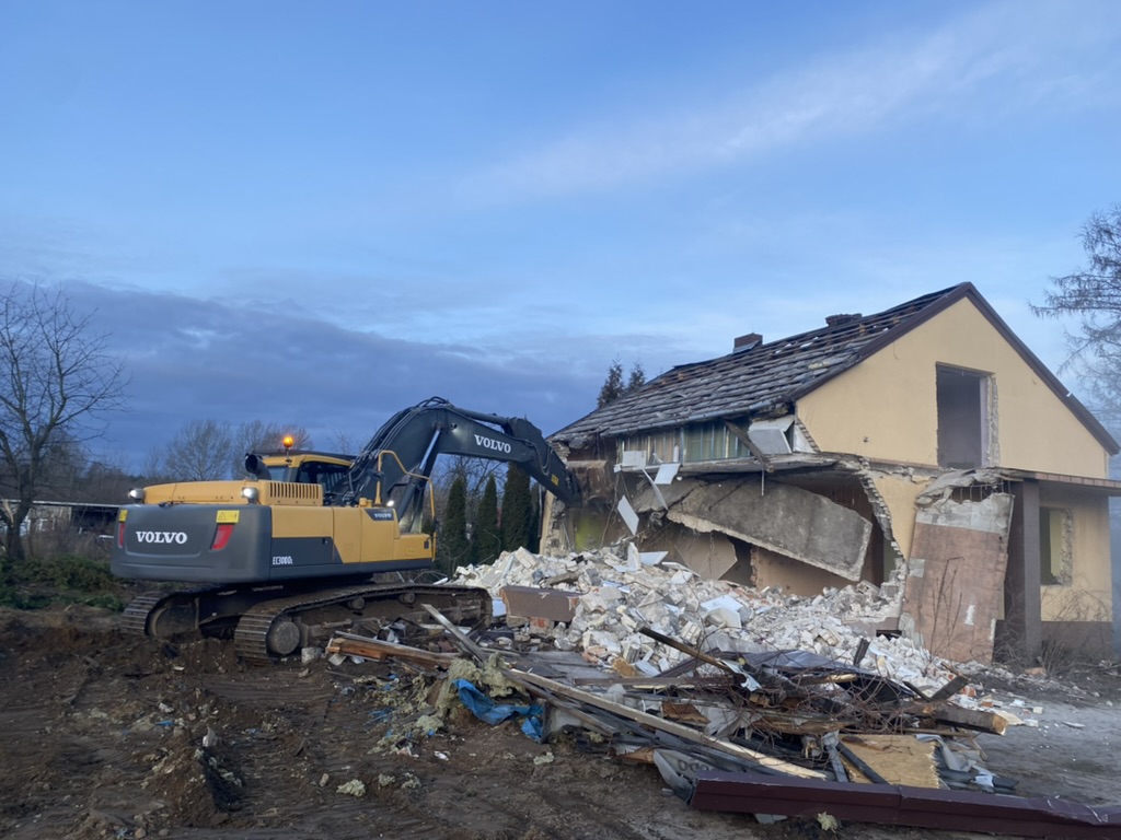 Yellow Volvo excavator demolishing a partially collapsed house with debris scattered nearby.