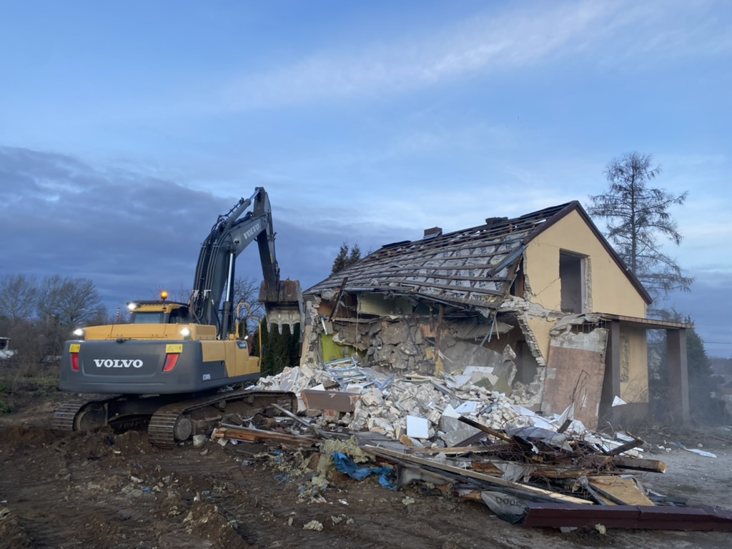 Yellow Volvo excavator demolishing a partially destroyed house with debris scattered around.