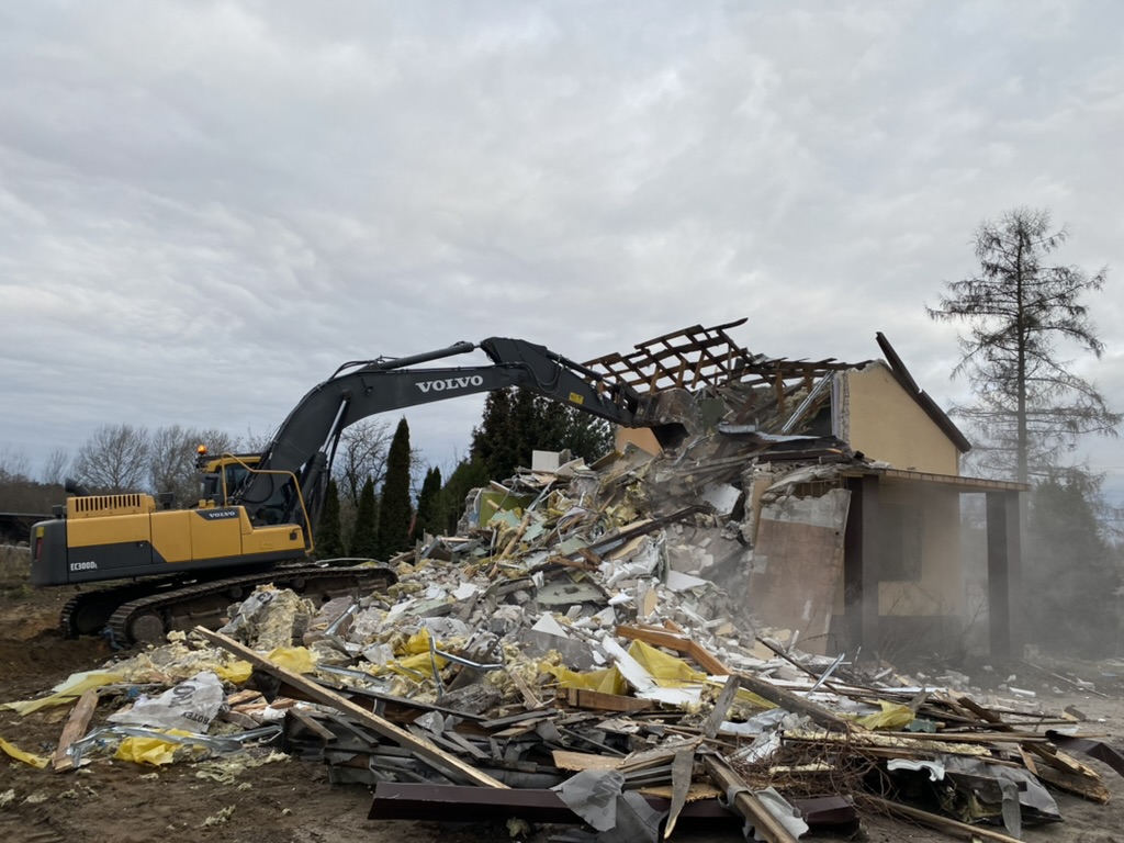 Yellow and black Volvo excavator demolishing a partially collapsed house with debris piled around it.
