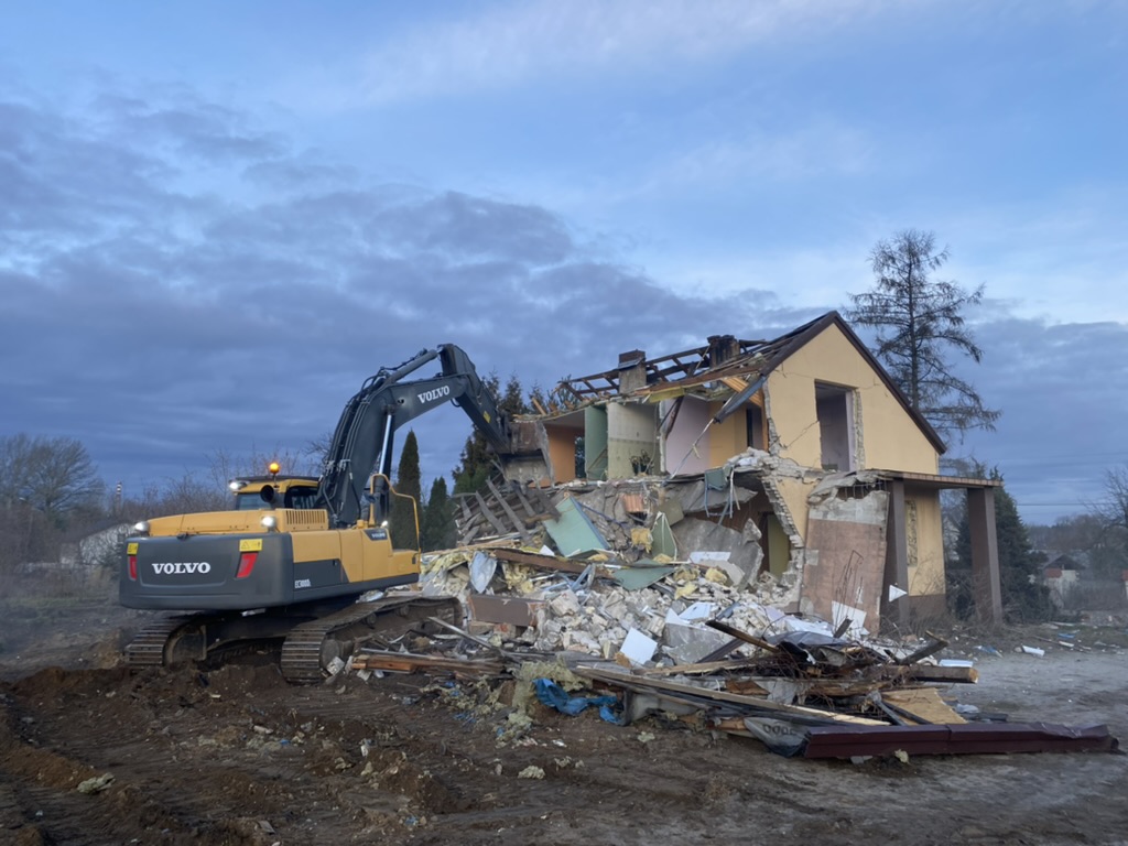 Yellow Volvo excavator demolishing a partially destroyed two-story house under a cloudy sky.