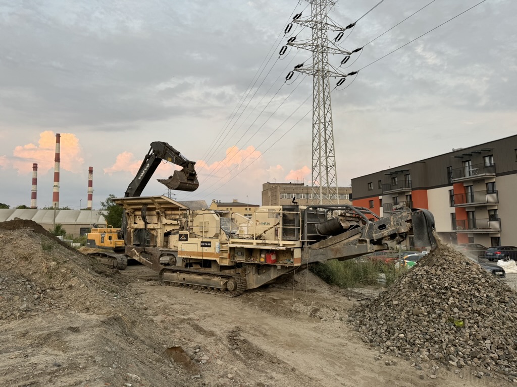 Construction site with a large yellow excavator loading rubble into a crusher machine near buildings.