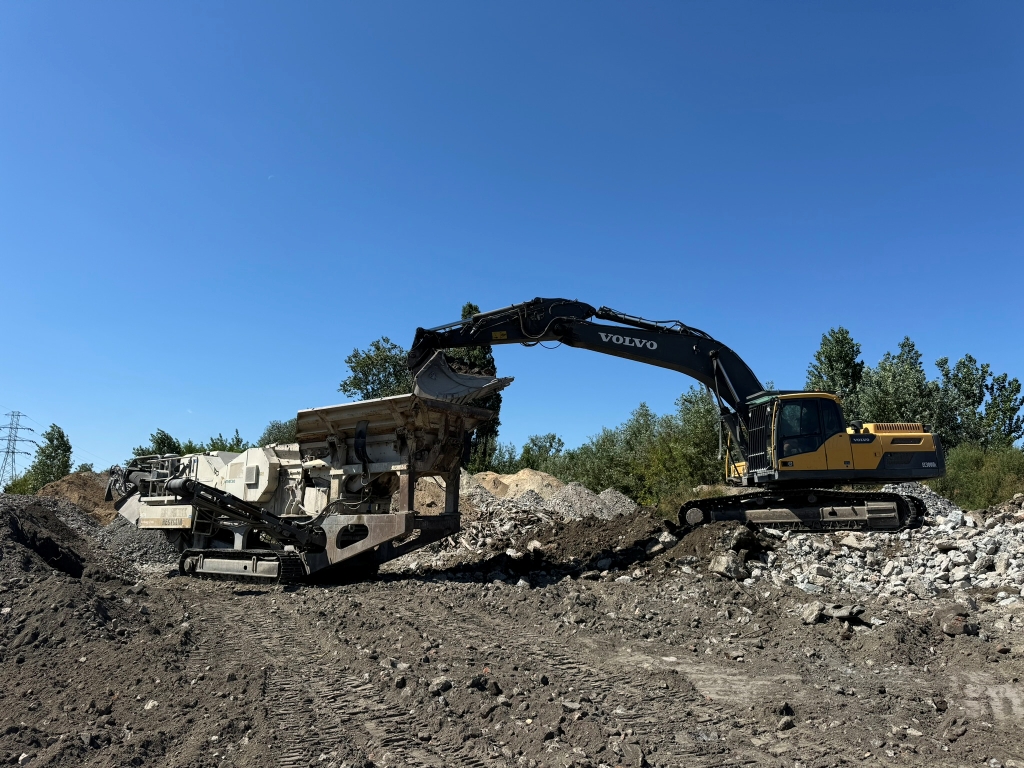 Yellow Volvo excavator loading dirt into a large crusher on a rocky construction site under a clear sky.