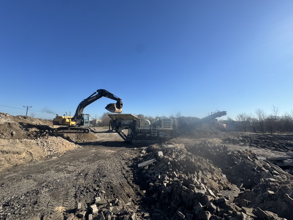 Excavator loading dirt into machinery at a construction site with piles of rubble under blue sky.