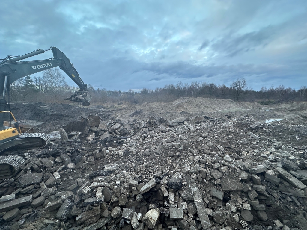 Excavator loading rubble and broken concrete in an open construction site with cloudy sky.