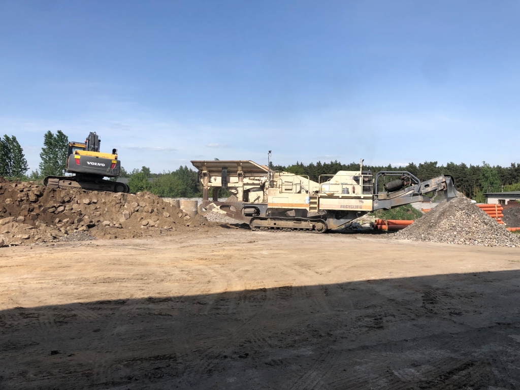 Construction site with yellow Volvo excavator on dirt pile and a recycling machine stacking gravel.