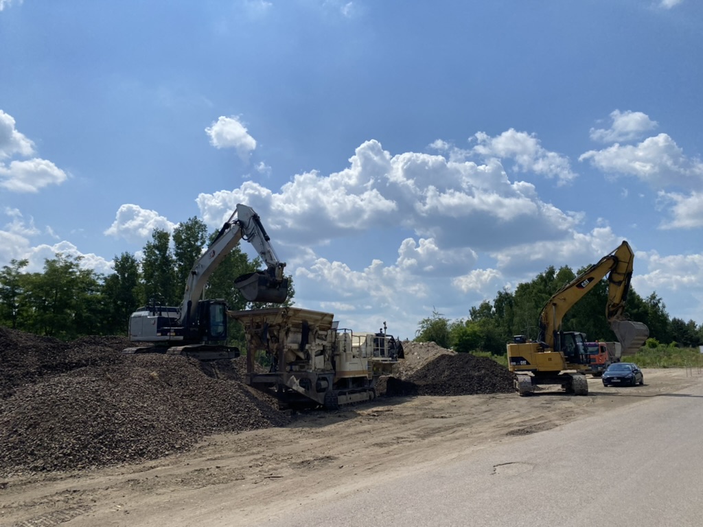 Two excavators working on large piles of dirt or gravel under a blue sky with scattered clouds.