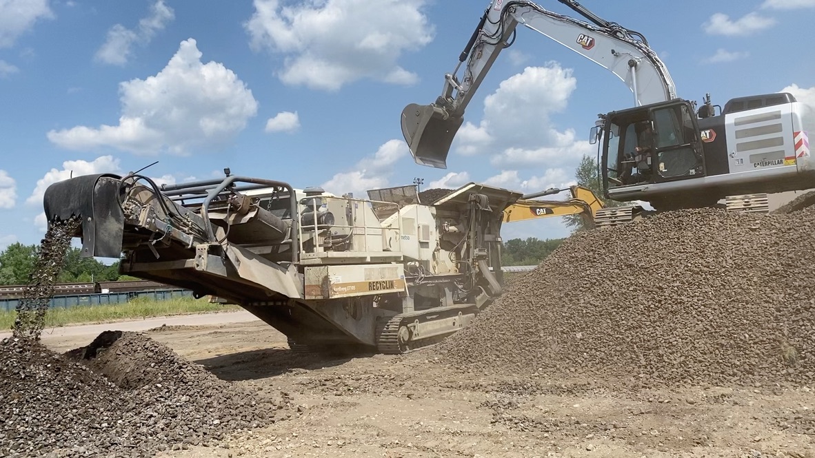 Excavator loading dirt into machine that sorts and deposits rocks into piles outdoors under a blue sky.