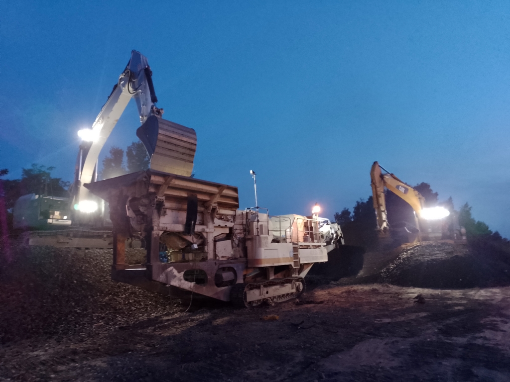 Two excavators loading material into a large crusher machine at dusk with their lights on.