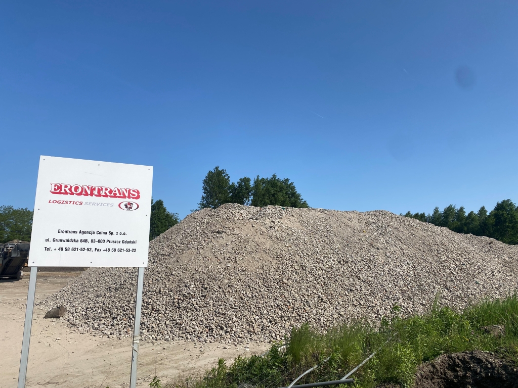 Large pile of gravel at an outdoor site with an Erontrans Logistics Services sign nearby.