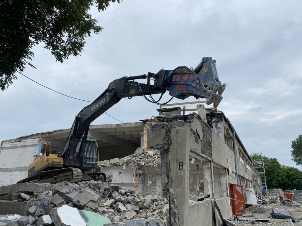 Volvo excavator demolishing concrete building with rubble piled around under cloudy sky.