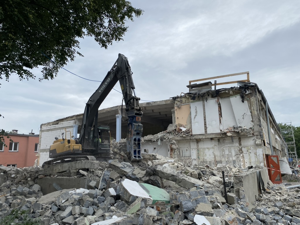 Heavy excavator demolishing a partially torn-down building with rubble piled in front under a cloudy sky.