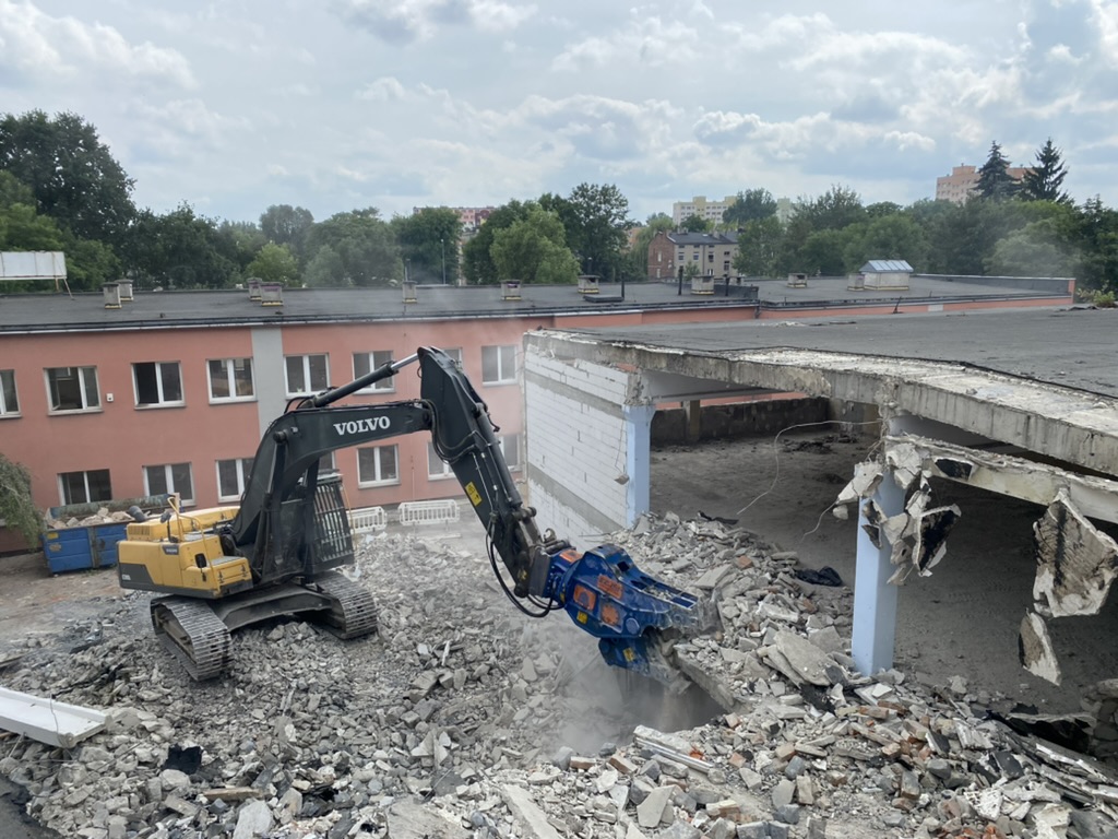 Black and yellow Volvo excavator demolishing concrete rubble beside broken building walls.