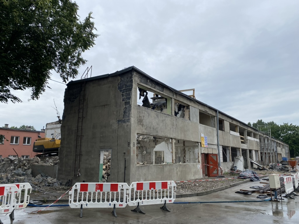 Partially demolished two-story building with construction barriers and equipment around it on a cloudy day.