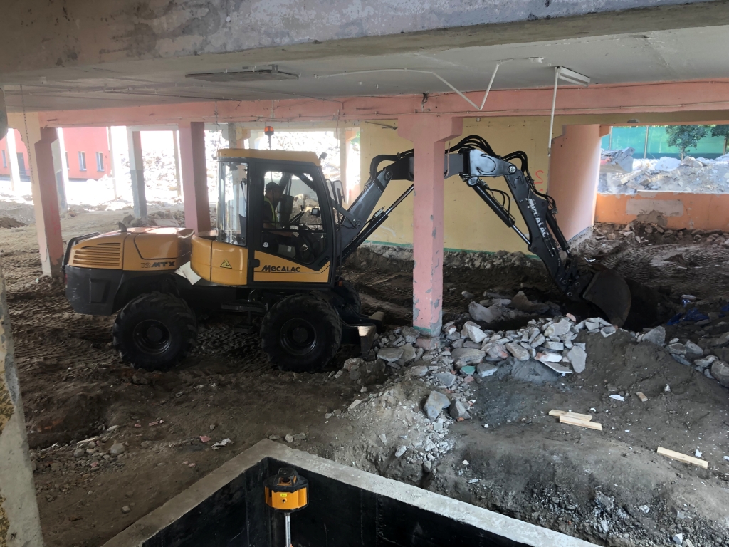 Yellow Mecalac excavator working inside a partially demolished building on dirt and rubble.