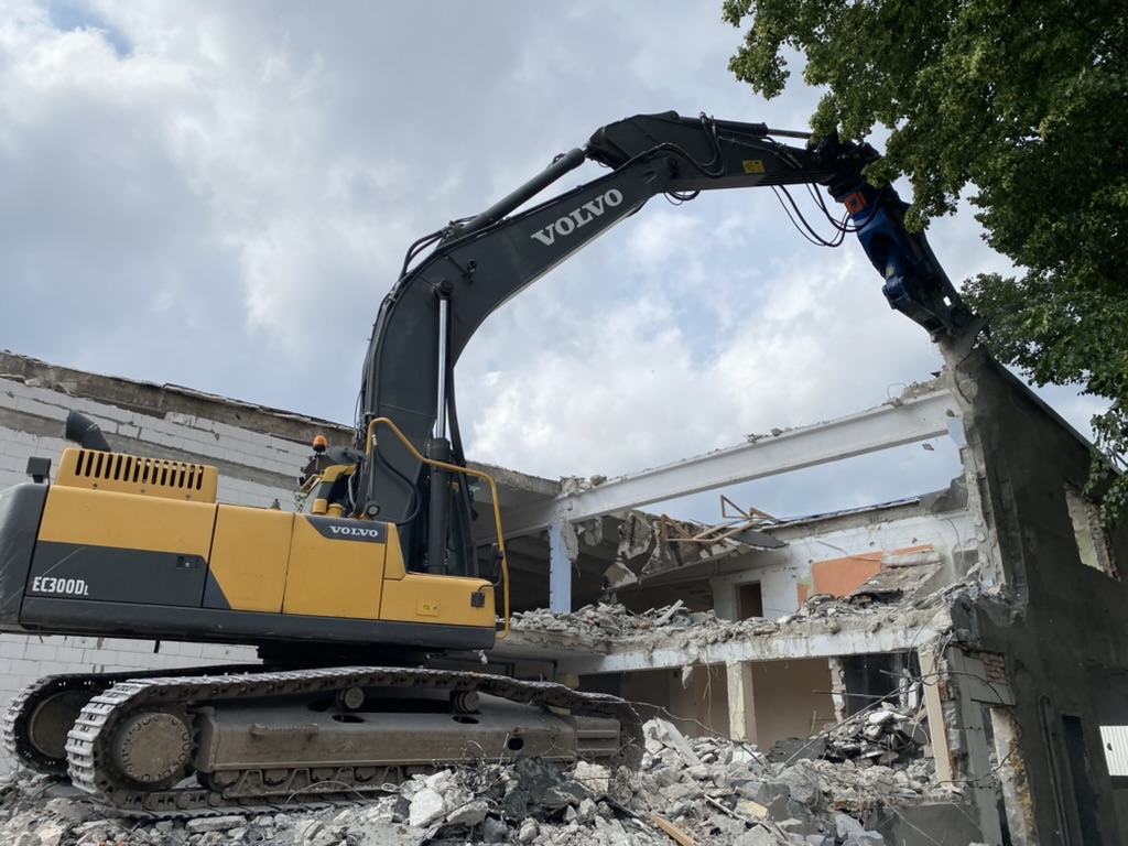 Volvo excavator demolishing a building with rubble on the ground and trees nearby.