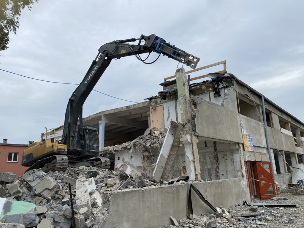 Excavator demolishing a partially destroyed concrete building amid rubble under a cloudy sky.
