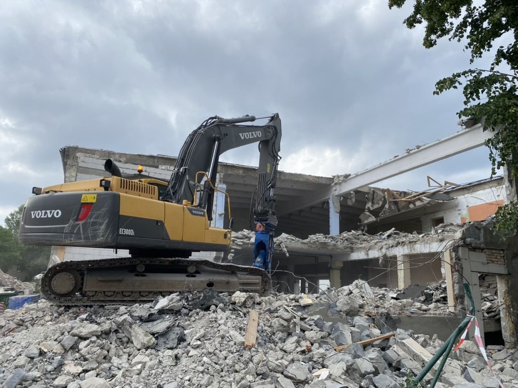 Yellow Volvo excavator stands on rubble at a partially demolished building under cloudy sky.