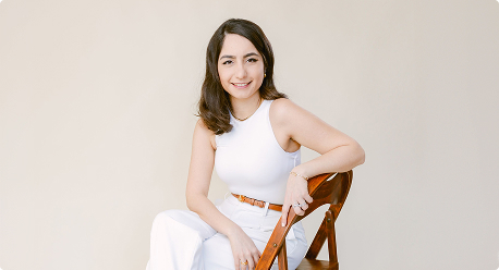 Jeune femme souriante assise sur une chaise en bois, vêtue de blanc, sur fond beige.