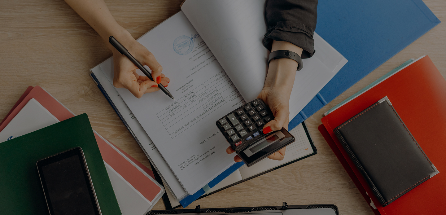 Mains tenant un stylo et une calculatrice au-dessus de dossiers et documents sur un bureau en bois.