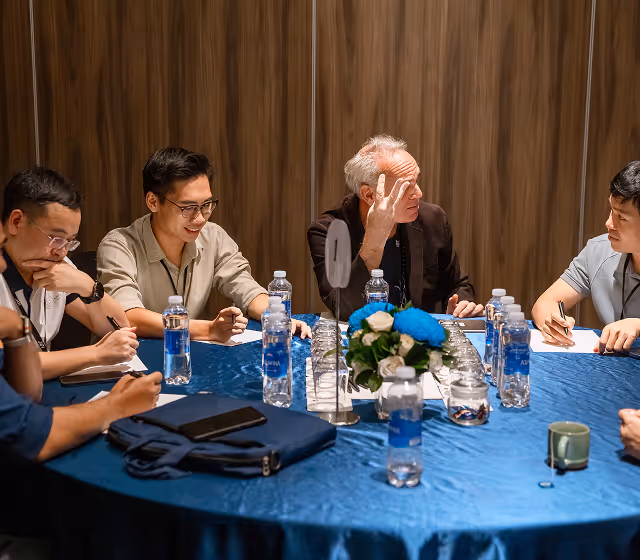 A group of men sitting around a table with blue tablecloth, engaged in discussion with water bottles and a floral centerpiece in the middle.