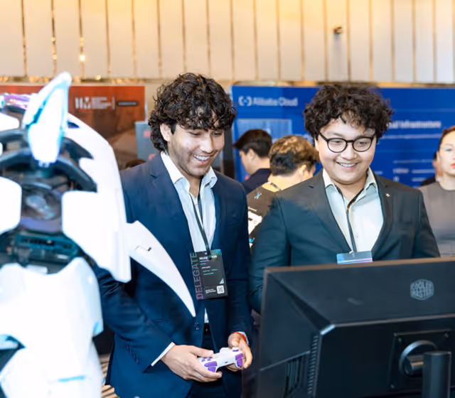 Two men in suits smiling and playing a video game with a controller next to a white robot figure.