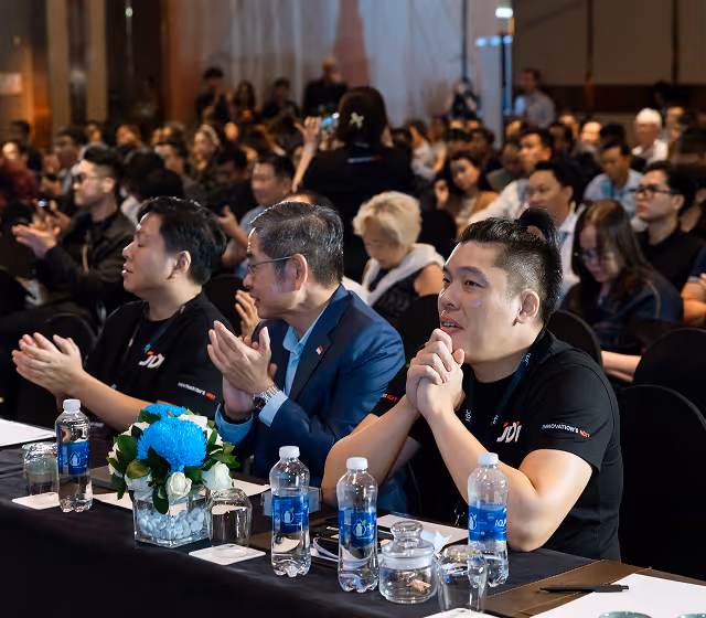 Three men sitting at a table with bottled water and flowers, attentively watching and clapping during an indoor event with a large audience.