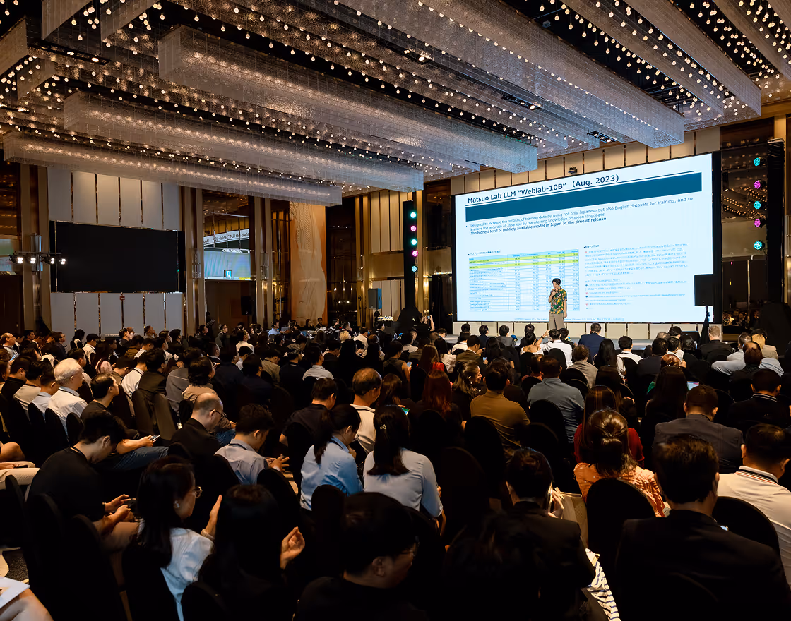 Two men in suits smiling and playing a video game with a controller next to a white robot figure.